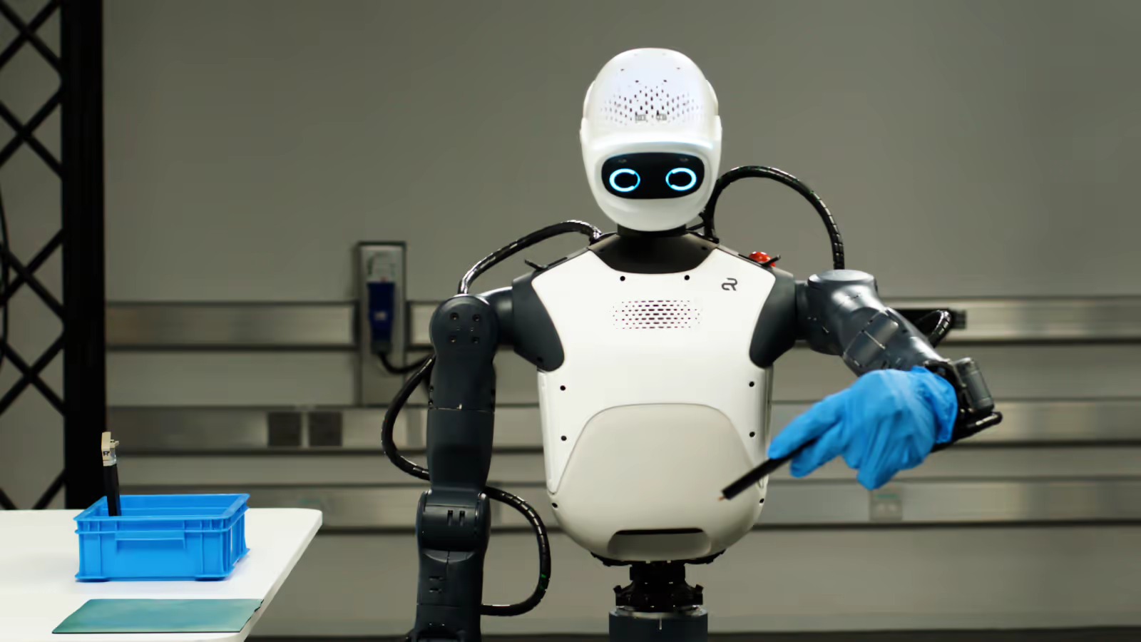 A white humanoid robot with glowing blue eyes and a blue glove pointing at a tool tray, from NVIDIA's October 2026 Isaac platform announcement.