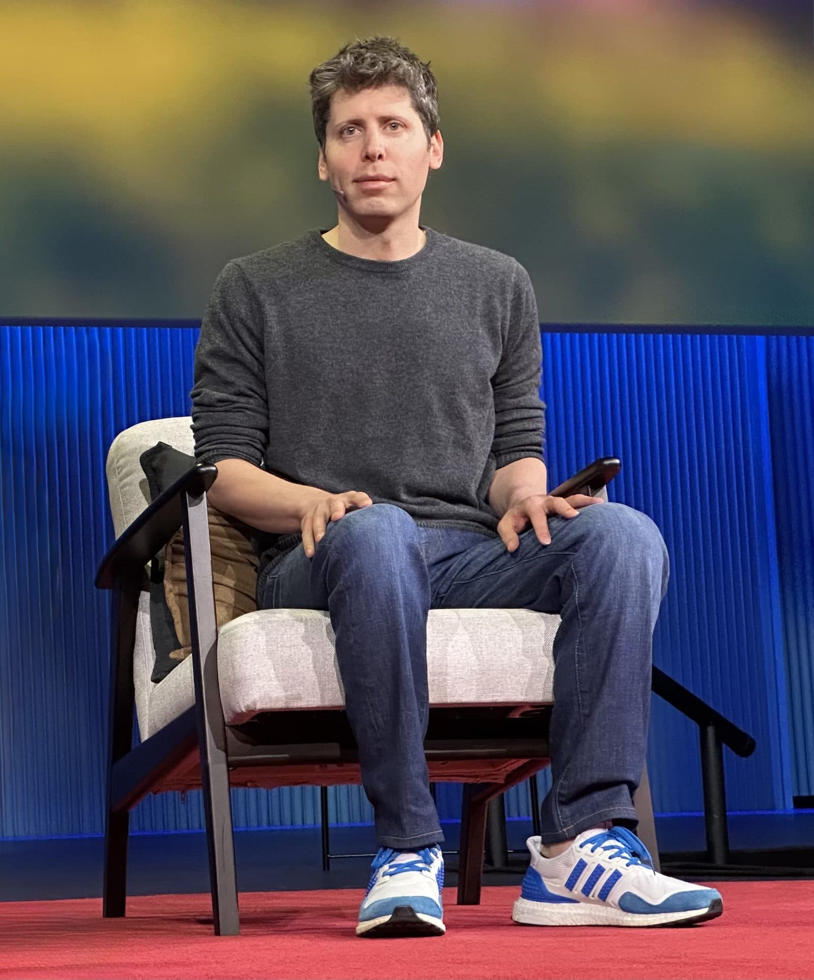 Sam Altman speaking on stage in a grey jumper and jeans, seated in an armchair with a lapel microphone, at TED in April 2025.