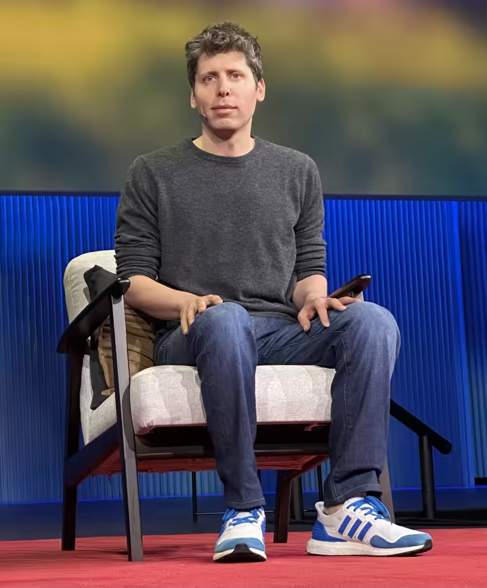Sam Altman seated on a stage in a grey jumper and jeans in front of a blue backdrop.
