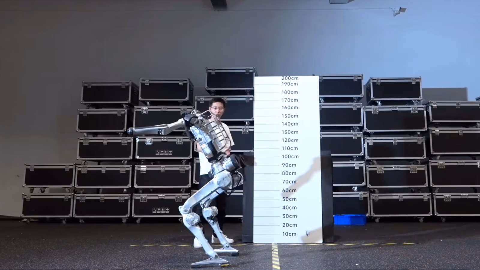 A Unitree humanoid robot demonstrator, filmed for the Superman reveal, standing with one leg raised beside a metre-marked height chart in a warehouse space.
