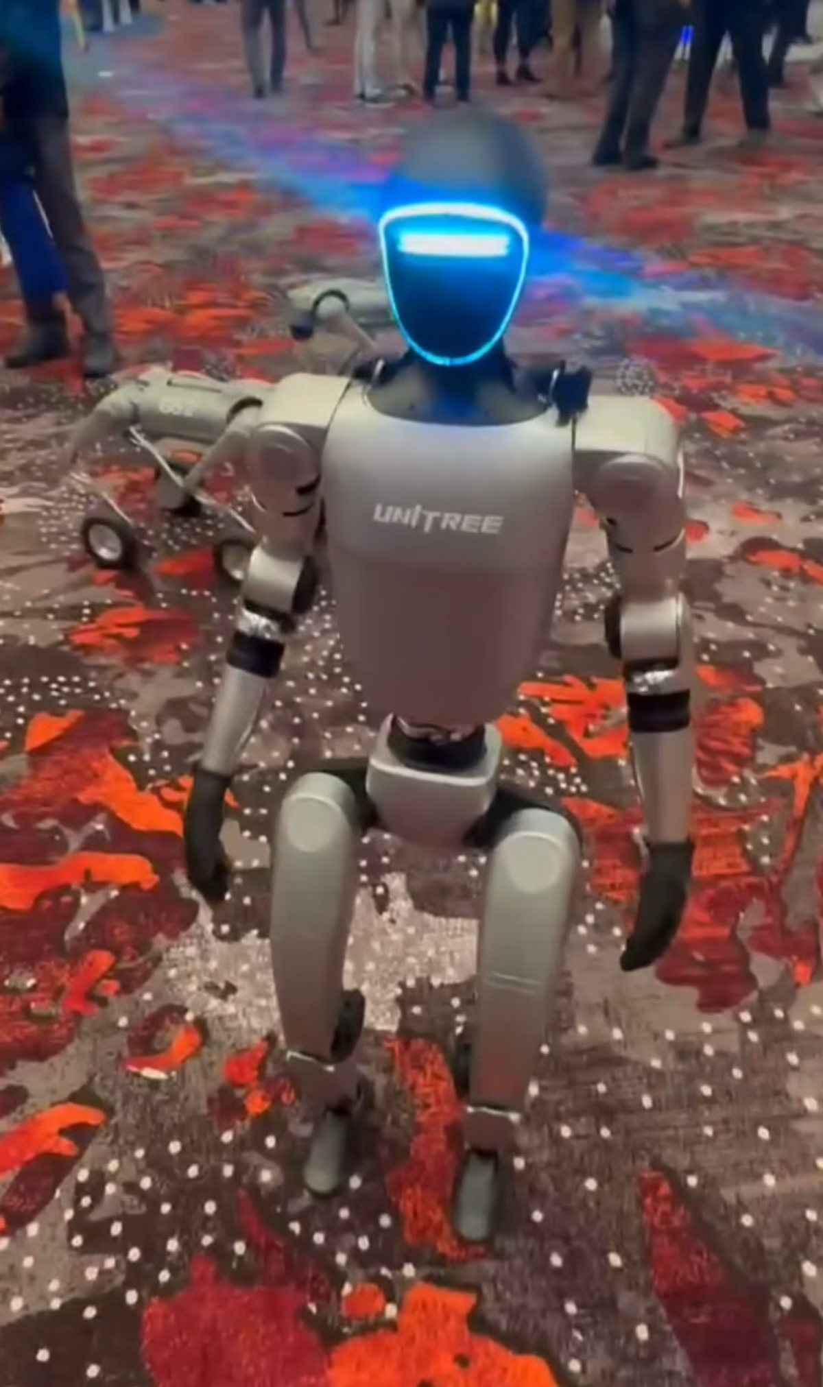 A Unitree G1 humanoid robot with 'Unitree' branding on its chest and a glowing blue face visor, standing on a patterned carpet at an indoor event.